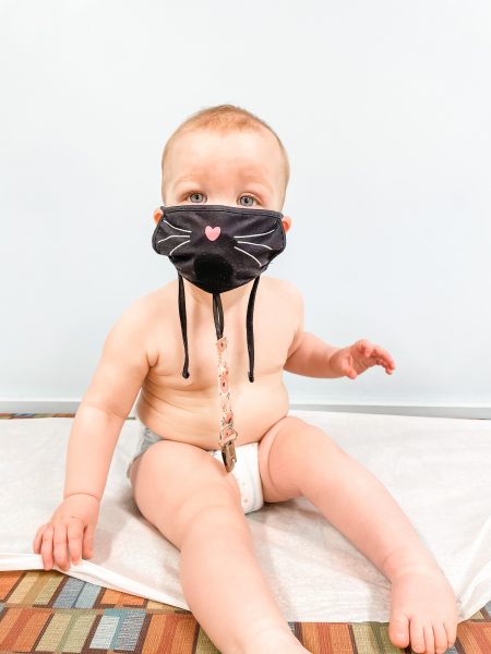 A diapered baby on an exam table with a black kitten face mask on